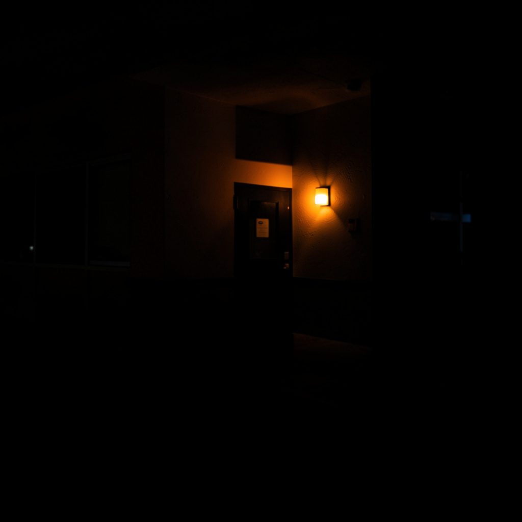 Dark low-key photograph of a quiet hotel desk with a softly glowing lamp, deep shadows, and warm muted browns.