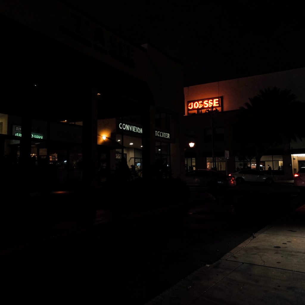Exterior of a low-rise hotel at dusk with warm porch lighting and quiet street, deep shadows and muted beige tones