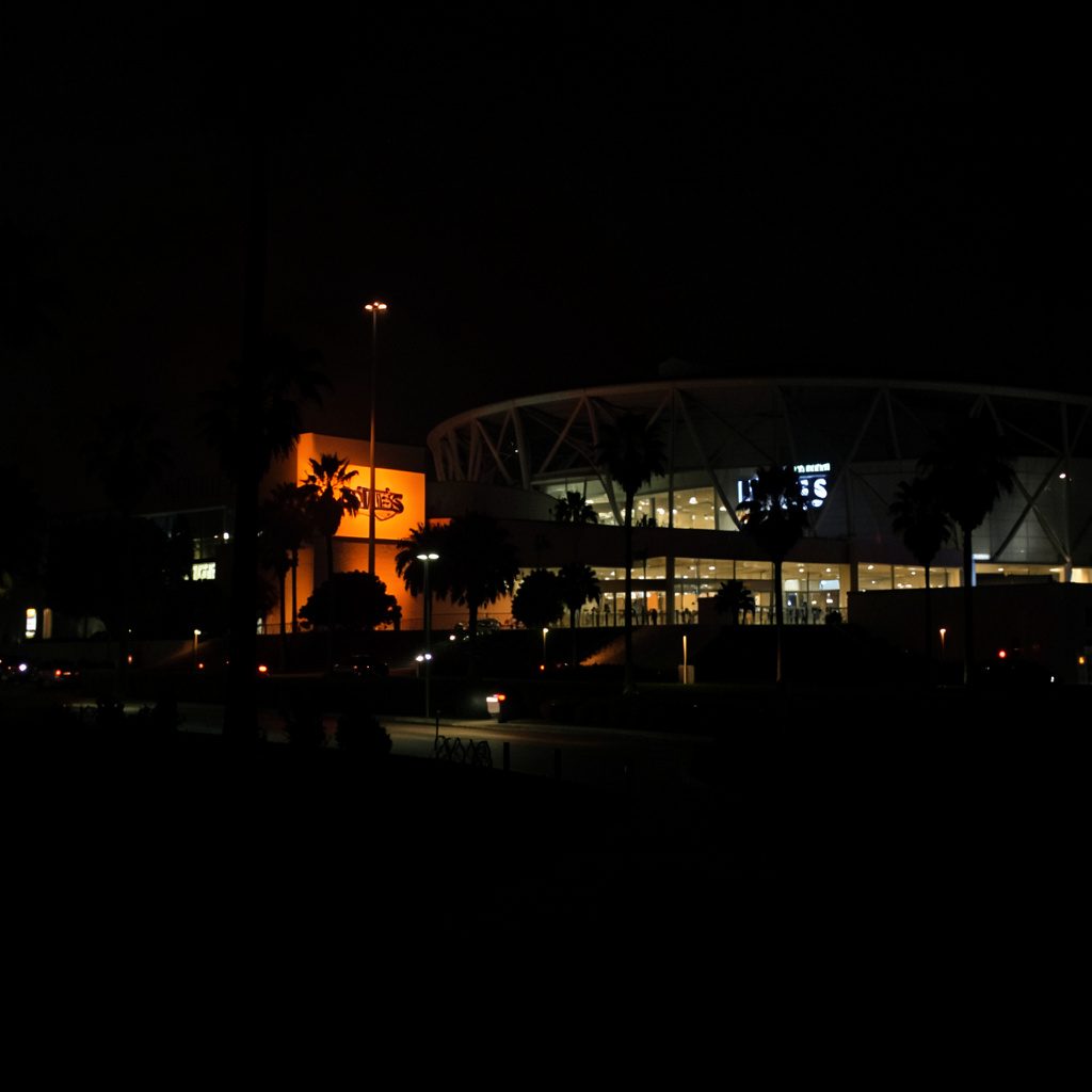 Dark, low-key photograph of a wide roadway near a California stadium at twilight with muted slate tones and soft glow of distant lights