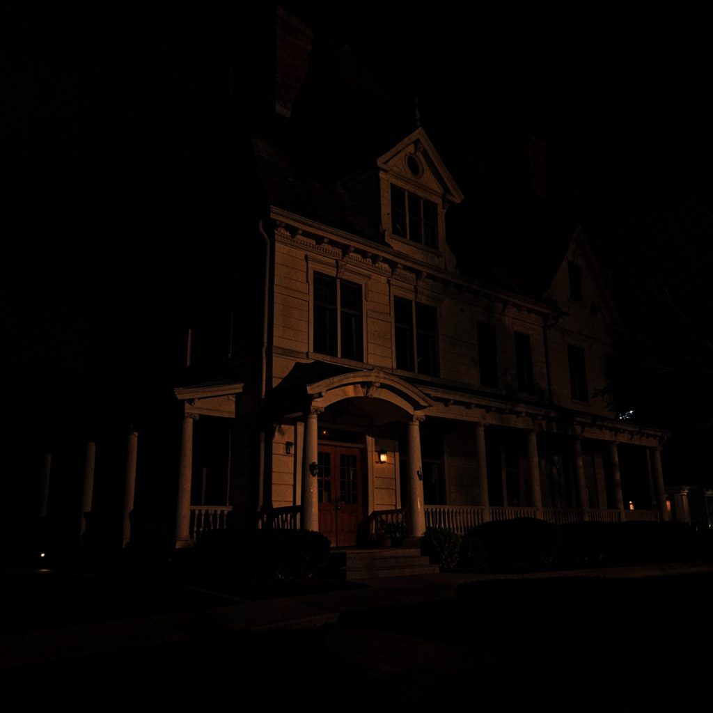 A dark, moody photograph of a large ornate Victorian-style mansion with turrets and staircases, shadowed under a deep blue-grey evening sky